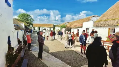 a group of people stand looking at a number of replica Irish famine era buildings. The buildings are painted white and have thatched roofs.