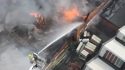 West Midlands Fire Service A firefighter on a platform fires a jet into the fire below as flames leap from the building. The flames are at the edge of the building with large items on the other side of a wall. There is no roof on the building. Burnt debris can be seen.
