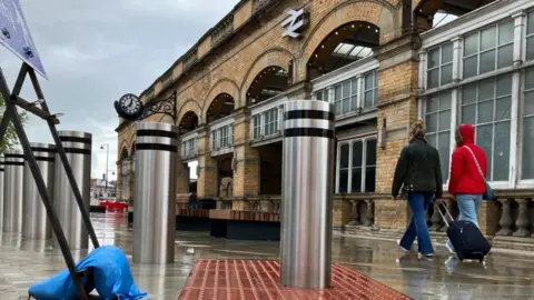 A bollard rises out of red tactile paving, with York Railway Station in the background.