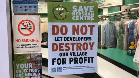 BBC Poster from campaign group Save Hersham Centre in the window of shop. The green and white poster has red, black and white writing and says: "Do not let developers destroy our village for profit."