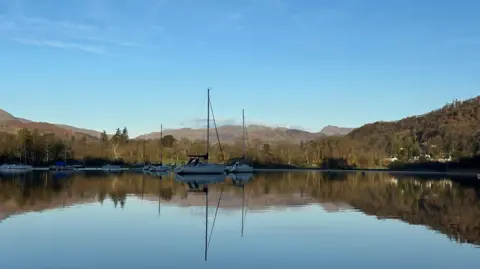 Mrs M View of the head of a lake with a number of small sailing boats - three with large masts - which are reflected in the still waters. The lake is fringed with trees with mountains in the distance.