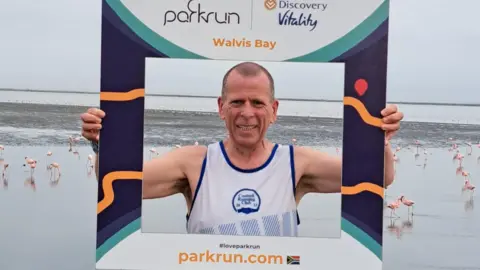 A runner in a white vest is holding up a cardboard photo frame that shows he is in Walvis Bay in Namibia. He is standing in front of a tidal lagoon and there are dozens of pink flamingos on the sand behind him
