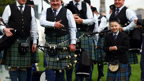 Getty Images A group of pipers wearing kilts standing next to young girls smiling as she also wears a kilt.
