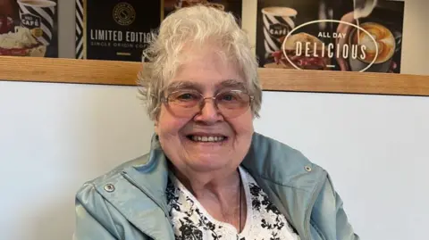 A woman with a grey coat and a white top underneath and who is wearing glasses is looking at the camera.