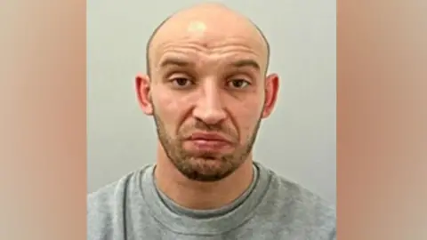 Slawomir Klimek's custody mugshot. A bald manwith brown eyes and dark stubble, he is wearing a grey sweatshirt.