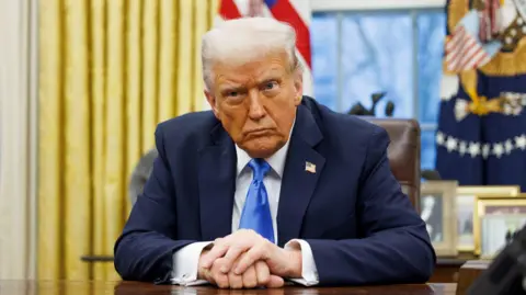 Donald Trump sits at the desk in the Oval Office. He is wearing a navy suit jacket, a white collared shirt and a blue tie. There is a US flag pin on his lapel.