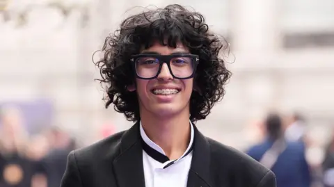 Getty Images Moosa Mostafa with a huge mop of curly hair, large glasses and braces on his teeth smiles at the camera. He is wearing a suit jacket and white shirt with a black grandad collar.