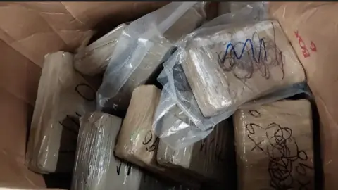 Cumbria Constabulary Several rectangular packages with brown tape and scribbled black writing on, in a box. 