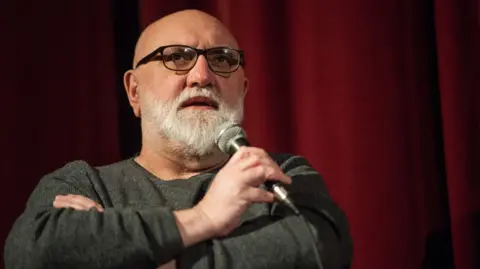 Guy Smallman/Getty Images Sayle, who has a white beard and no hair, is holding a microphone and is in front of a red curtain. He is wearing glasses and a grey top.