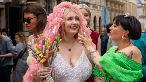 Contributed A smiling woman in a bridal dress holds a bunch of colourful flowers as a friend in a green top helps adjust a pink head decoration. In the background other well wishers can be seen