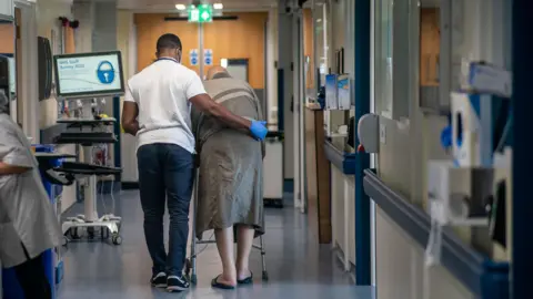 A male nurse supports a male patient as they walk down a hospital corridor, surrounded by hospital equipment