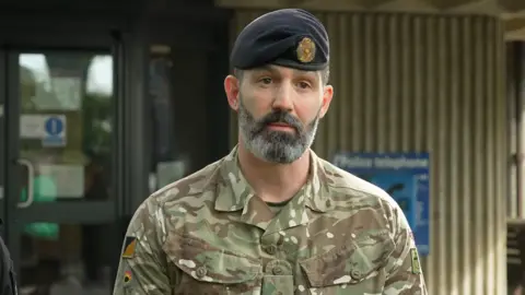 The image shows a single adult wearing a camouflage military uniform and a dark beret with an insignia, photographed from the chest up outdoors. 