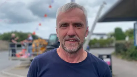 BBC Peadar is looking directly into the camera wearing a navy blue t-shirt. The background of the photo is blurred but shows men erecting orange and white bunting.