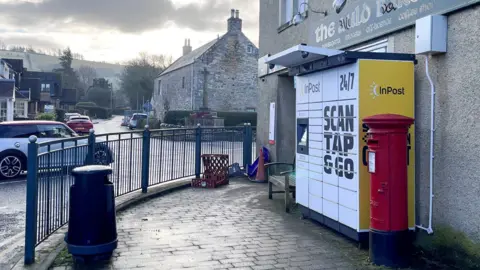 An InPost locker outside a shop in the Borders on a grey day with a car driving past
