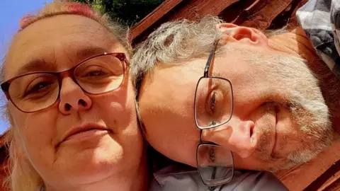 Handout A woman in glasses looks into the camera, light hair tied back. The top has pink streaks in. A man in glasses rests his head on her shoulder, he also wears glasses and looks at the camera with a small smile. What looks like a wooden fence is behind them, a patch of blue sky and green tree visible over the top. 