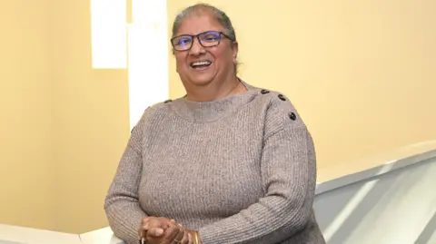 Mr Kaur stands against a pale yellow backdrop, smiling. She has grey hair worn up, and wears black-framed glasses and a beige knitted jumper with tortoiseshell buttons along the shoulders.