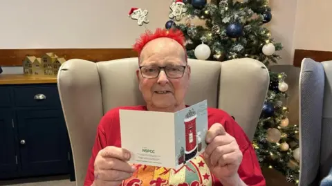 The image shows Peter sitting in a cushioned armchair, holding an open greeting card. The card features a red postbox with snow on top. Peter is wearing a bright red shirt with a festive design and has a red headband decorated with two small gingerbread men. In the background, there is a decorated Christmas tree adorned with silver and white baubles. To the left, there is a dark blue cabinet with small decorative wooden houses placed on top, adding to the cozy holiday setting.