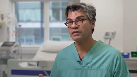 Ahmed Ahmed sat in a hospital room. Behind him is a hospital bed and clinical machines. He is wearing green scrubs and has on glasses.