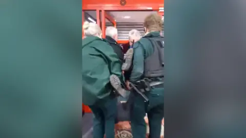 Officers in uniform and a man carry a woman out of a shop. Her legs and feet are being held up by the officers who are carrying her. Shutters are partially down at the doors of the shop.