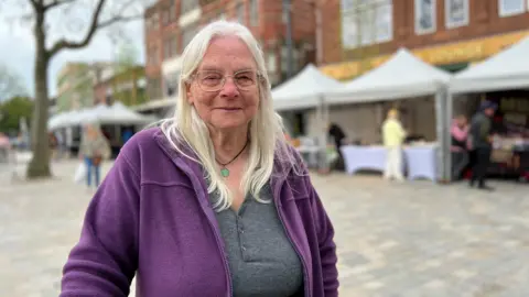 Fen Watson is in the middle of Carlisle. She has long hair, glasses and is wearing a purple jacket. There are market stalls behind her.