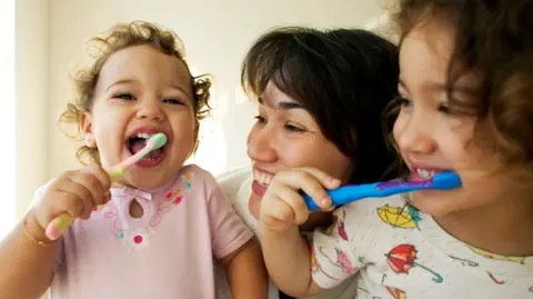 Getty Images A woman smiles at two grinning children brushing their teeth