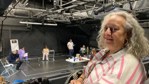 A woman with grey, curly hair and pink and white-striped top in the foreground with actors in a rehearsal in the background.