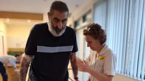 Anthony Dexter with Marion Packwood. Anthony, has short dark hair and a grey heard, is wearing a black T-shirt, and Marion, is on hand to his right. They are in a rehab centre. 