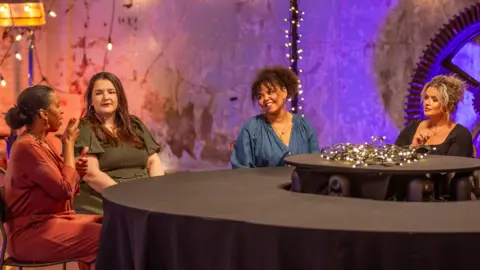 Becky Bailey/LumoTV Four women are sitting around a table signing to each other. One woman is wearing a coral outfit, another a green dress, another a blue top and another a black t-shirt. 