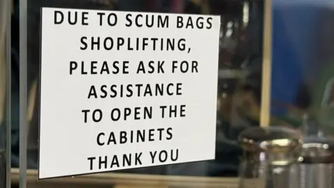 Rob Davies' printed signs showing 'Due to scum bags shoplifting, please ask for assistance to open the cabinets, thank you'