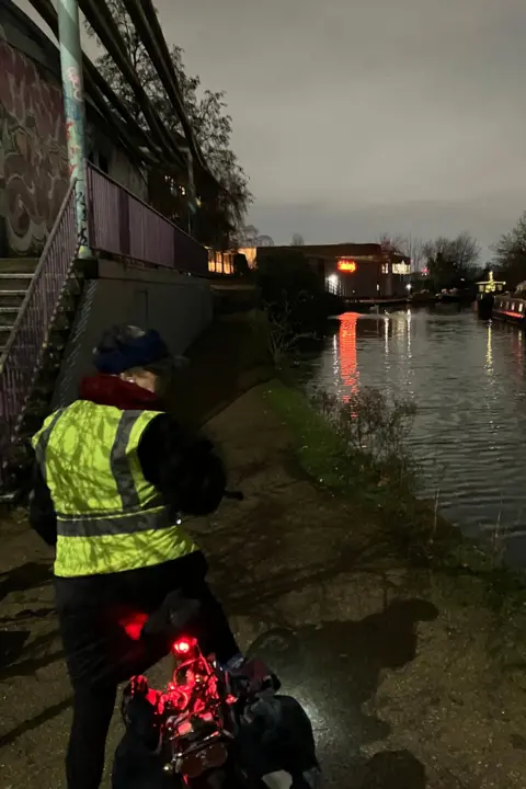 LCC Back view of female cyclist paused at the edge of a canal. She wears yellow hi-vis jacket and her red rear cycle light glows in the dusk.