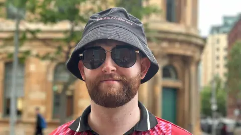 A young man with a ginger beard wearing a black bucket hat, sunglasses and a red and black collared top.