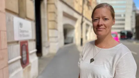 BBC/ Hazel Shearing Rebecca Larkin usa uma camiseta branca com um colar de prata. É um dia ensolarado e ela está na rua em frente ao Departamento de Educação de Londres.