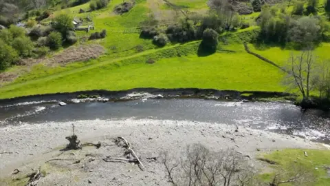 A drone shot of the River Elwy, there is a completely dry grey riverbed next to the river while is now flowing into a nearby green field. In this birds eye view shot there also trees and a couple small buildings. 
