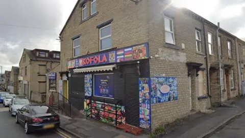 The exterior of Kofola in Great Horton Road, Bradford. The shop has a blue and red sign as well as pictures of food items on its brick walls. There are cars parked in front of the shop.