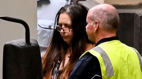 Getty Images A woman with long brown hair, wearing glasses, stands between a vehicle and a man in a vest with high visibility 