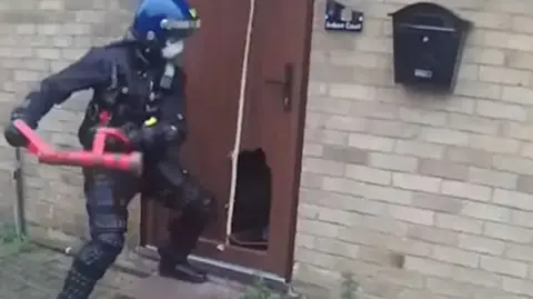 Cambridgeshire Police A police officer with a red smashing their way into a brown door at a brick house