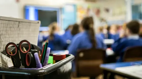 File photo showing children in blue jumpers sitting at school desks. They are all blurry in the background, in the foreground is a box containing pencils and other school stationery.
