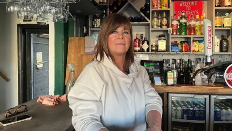 Tony Fisher/BBC Louise Maloney sitting behind the bar in her pub. Wine glasses hang down from the bar above and there are bottles of spirits and beers behind her, as well as two fridges full of cans. Louise looks at the camera with a half smile, she wears a white hooded top and has shoulder length brown hair with a fringe.