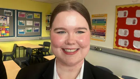 Meghan, a teenage schoolgirl, smiles at the camera while standing in a classroom with yellow walls. She has blue eyes and straight, light brown hair tied back from her face. She is wearing a white shirt and a black blazer. There are desks and chairs in the background and there are educational posters on the walls. 