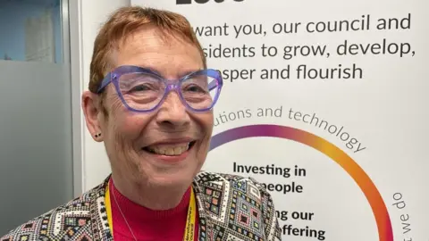 Conservative Councillor Carole Gandy, the cabinet member for adult social care at Herefordshire Council, is smiling at the camera - she is wearing a patterned jacket and a red jumper.