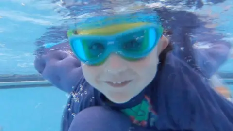 A toddler is underwater in a swimming pool smiling. They are wearing a pair of swimming goggles and a cape is billowing behind them.