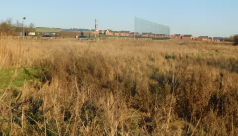 LDRS An area of land covered in long grass with a sports ground and houses to the rear