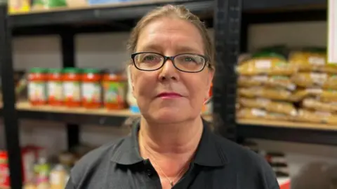 Laura Foster/BBC A woman with tied back hair wearing dark-framed glasses and a black top standing in front of shelves of food