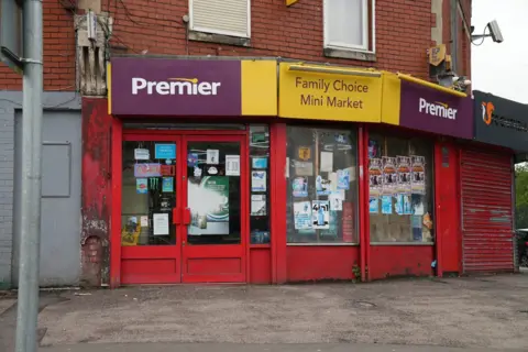 The outside of a Premier shop. Which is painted red, with the purple and yellow Premier branding above and Family Choice Mini Market written above the door. There are numerous posters stuck on the windows, and a pavement in front.