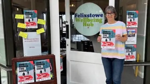 Karen Goddard Karen stands in front of the Felixstowe Wellbeing Hub. The windows are covered in posters for her play Don't Knock the Dockers. Karen is wearing a blue, yellow and pink striped T-shirt and jeans. She has glasses and bobbed dark grey hair