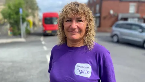 Tina Leslie, a woman with long, curly blonde hair, is stood on a street and is smiling at the camera. She is wearing a purple T-shirt branded "freedom4girls".