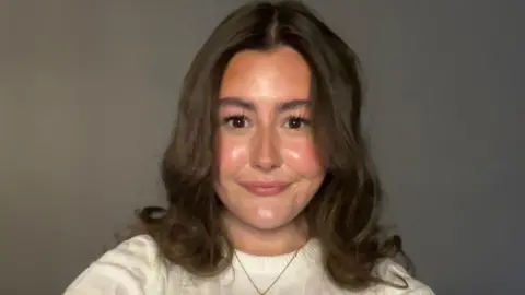 Aoife, a 16-year-old girl, taking a selfie. She is wearing a white knitted top and has brown, curly hair that goes to her shoulders. She is wearing a gold chain around her neck and is in front of a neutral background