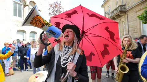 Derry City and Strabane District Council A woman holding a megaphone and umbrella is wearing a top hat. She is singing into the megaphone, a saxophone player stands to her right, while a crowd can be seen in the background.