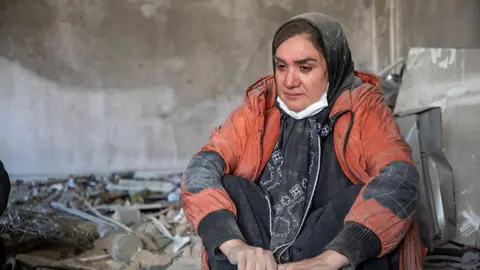 A woman sitting crying covered in dust as she sits surrounded by rubble
