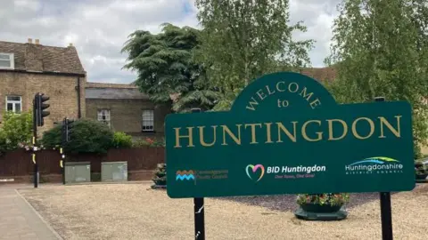 A green sign reading "Welcome to Huntingdon" with a gravel car park behind it and houses